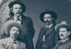 Black and white vintage photo of four men from the late 1800s, all wearing wide-brimmed hats and suits. Two are seated and two are standing behind them. The man on the left is wearing a light-colored suit and cowboy hat; the others are in darker suits. They have mustaches and long hair, styled in the fashion of the Old West. The background features an arched doorway.
