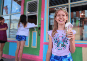 A smiling girl holding a large ice cream cone stands in front of a colorful storefront with pink, green, and blue trim. Two other children are near a walk-up service window in the background.