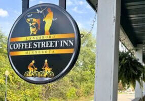Round black-and-gold Coffee Street Inn sign in Lanesboro, Minnesota, featuring graphics of a fisherman and cyclists, hanging outside the inn against a backdrop of trees and blue sky.