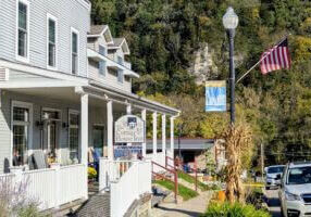 The Cottage House Inn in downtown Lanesboro, Minnesota, decorated with fall touches including cornstalks, pumpkins, and flowers along the sidewalk, with bluffs covered in autumn trees in the background.