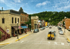 Downtown Lanesboro, MN with historic buildings, American flags, and a truck hauling kayaks and tubes—set against lush green bluff country under a blue sky. Photo by Explore Minnesota / Ryan Taylor.