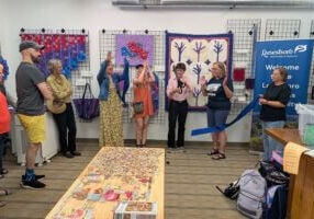 Community members and leaders gather inside Driftless Fiber Arts Collective for a ribbon cutting ceremony, surrounded by colorful fiber art and quilts on display.