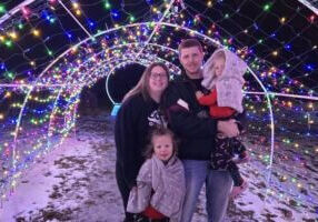 A family stands together beneath a colorful tunnel of holiday lights at the Lanesboro Legion Lights display on a winter night.