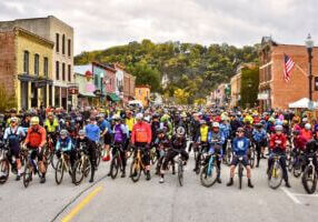 Hundreds of cyclists line up on Lanesboro’s historic downtown street for the start of the Filthy 50 Gravel Bike Race, surrounded by colorful storefronts and autumn bluff scenery.