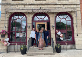 Five people stand on the front steps of the historic Hotel Lanesboro in downtown Lanesboro, Minnesota, framed by arched windows, stone façade, and welcoming entry.
