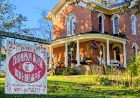 The James A. Thompson House Bed & Breakfast in Lanesboro, Minnesota—a beautifully restored 1870s Italianate brick mansion with white columns, arched windows, and a welcoming front porch decorated with pumpkins and fall flowers. A vintage-style sign in the foreground reads “James A. Thompson House Bed & Breakfast.”