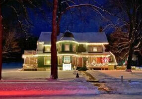 Historic Oakenwald Terrace mansion in Chatfield, Minnesota illuminated with warm white Christmas lights on a snowy winter evening, showcasing its Victorian architecture, wraparound porch, and festive holiday decorations under a dark blue sky.