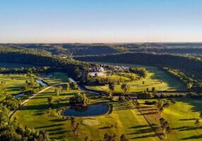 Aerial view of the Old Barn Resort and Rivers Bend Golf Course near Lanesboro, Minnesota, surrounded by rolling green hills, tree-lined fairways, a winding river, and a small pond under clear blue skies.