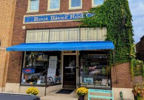 Front entrance of Root River Rod Co. in downtown Lanesboro, Minnesota, with a bright blue awning, ivy climbing the brick wall, and yellow mums by the doorway.