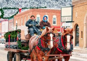 A horse-drawn carriage makes its way down Lanesboro’s snow-covered main street, driven by two warmly dressed locals. The decorated wagon, holiday wreaths, and twinkling lights create a festive small-town winter scene.