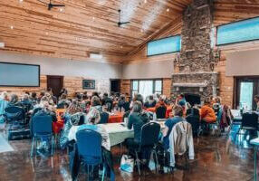 A group gathers inside a rustic wood-paneled event center with high ceilings, large windows, and a stone fireplace, attending a workshop or retreat in Lanesboro during winter.