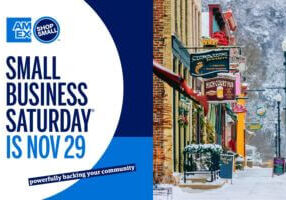 A promotional image for Small Business Saturday featuring the American Express Shop Small logo alongside a snowy winter street scene in downtown Lanesboro, Minnesota, with colorful storefronts and festive decorations.